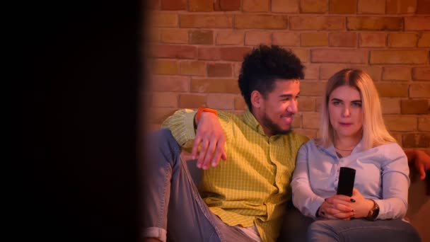 Dolly shot of young african guy with his blonde caucasian girlfriend watching TV in cozy home atmosphere. — Stock Footage Dolly shot of young african guy with his blonde caucasian girlfriend watching TV in cozy home atmosphere. Video Clip