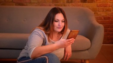 Rahat artı boyutu uzun saçlı model rahat ev atmosferinde katta oturan akıllı telefon içine sevinçle saatler.