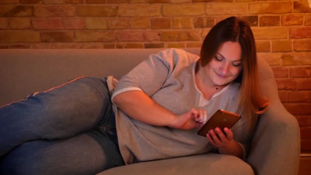 Portret van ontspannen plus grootte langharige model liggend op de Bank chatten vreugdevol op smartphone in gezellige huiselijke sfeer. — Stockvideo