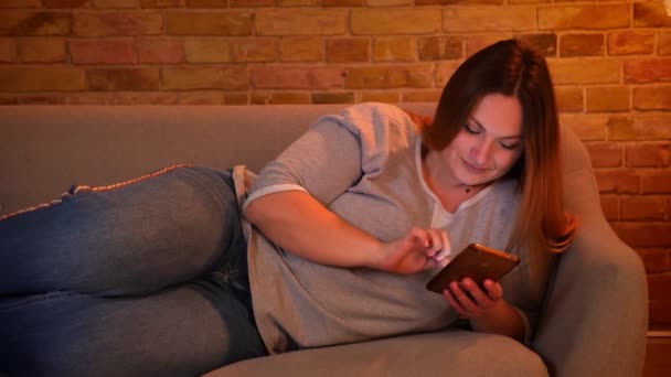 Porträtt av avslappnad plus size långhårig modell som ligger på soffan chatta glatt på smartphone i mysig hem atmosfär. — Stockvideo