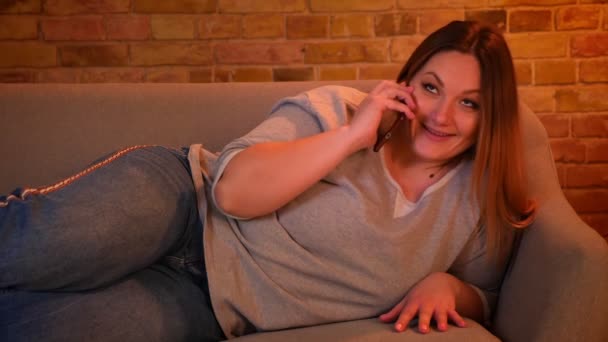 Портрет расслабленной модели с длинными волосами, лежащей на диване и говорящей по телефону в уютной домашней обстановке . — стоковое видео