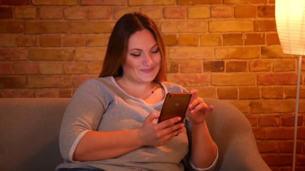 Retrato de relaxado plus size modelo de cabelos compridos assistindo em smartphone e rindo em ambiente acolhedor casa . — Vídeo de Stock