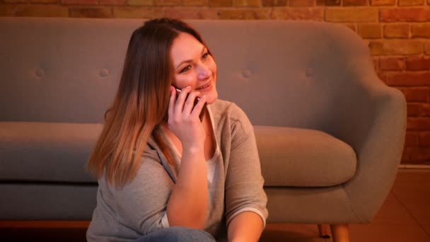 Πορτρέτο του χαλαρό συν μέγεθος μακριά μαλλιά μοντέλο κάθεται στο πάτωμα έχοντας μια φωνητική κλήση στο smartphone σε ζεστή ατμόσφαιρα στο σπίτι. — Αρχείο Βίντεο