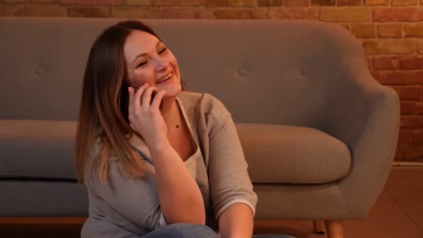 Συν μέγεθος μακριά μαλλιά μοντέλο κάθεται στο πάτωμα συνομιλίες χαρούμενα γελώντας στο smartphone σε ζεστή ατμόσφαιρα στο σπίτι. — Αρχείο Βίντεο