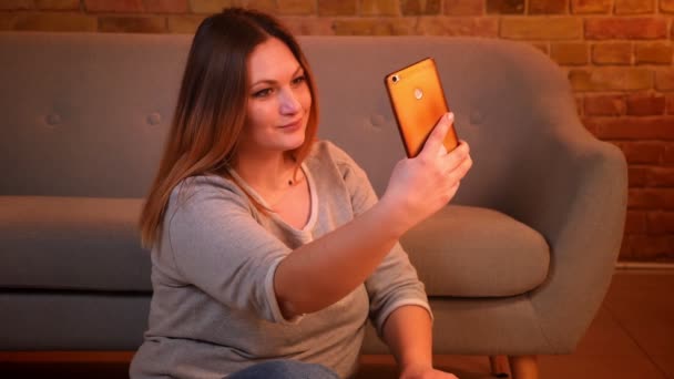 Modèle taille plus avec des cheveux longs faisant de jolies selfie-photos à l'aide d'un smartphone dans une atmosphère confortable à la maison . — Video