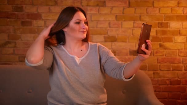 Modèle taille plus avec des cheveux longs faisant de jolies selfie-photos à l'aide d'un smartphone dans une atmosphère confortable à la maison . — Video