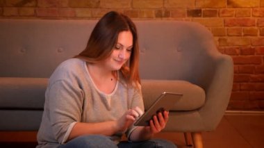 Kilolu uzun saçlı kadın serbest rahat ev atmosferinde tablet ile keyifli bir şekilde çalışan katta oturur.