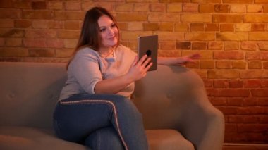 Kilolu uzun saçlı kadın model tablet bir videoarama sahip ve rahat ev atmosferinde gülüyor kanepeüzerinde oturur.