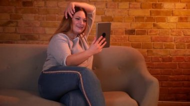 Kilolu neşeli kadın modeli rahat ev atmosferinde tablet bir videocall sahip kanepede oturur.