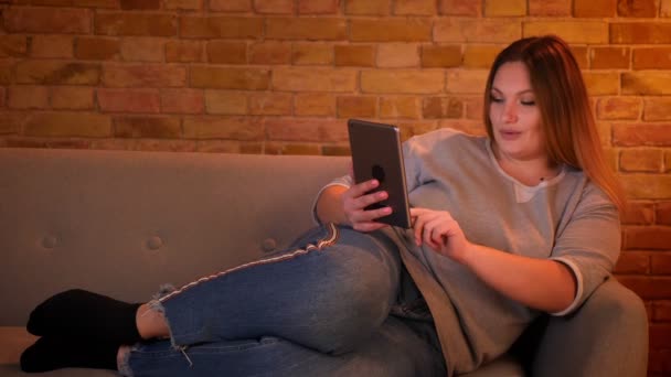 Porträtt av avslappnad plus size långhårig modell ligger på soffan chatta glatt på tablett i mysig hem atmosfär. — Stockvideo