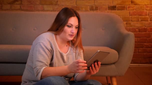 Sobrepeso de pelo largo freelancer femenino se sienta en el suelo trabajando con la tableta con atención en el acogedor ambiente del hogar . — Vídeo de stock