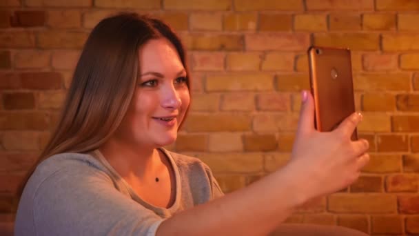 Modèle féminin de taille plus ayant un vidéocall sur smartphone et fixant ses cheveux joliment dans une atmosphère confortable à la maison . — Video