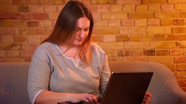 Sobrepeso de pelo largo freelancer femenino se sienta en el sofá trabajando con atención con el ordenador portátil en un ambiente acogedor en casa . — Vídeos de Stock