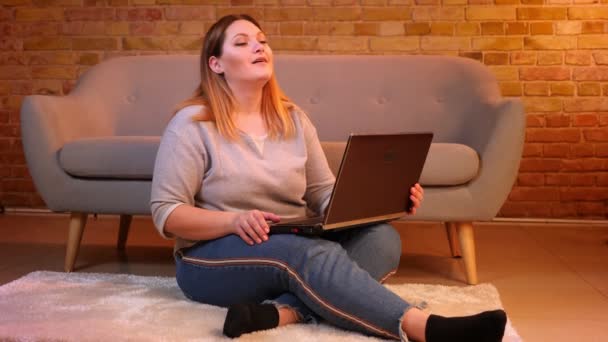 Além disso modelo tamanho senta-se no chão falando em videochat no laptop sendo pensativo e alegre em ambiente acolhedor casa . — Vídeo de Stock