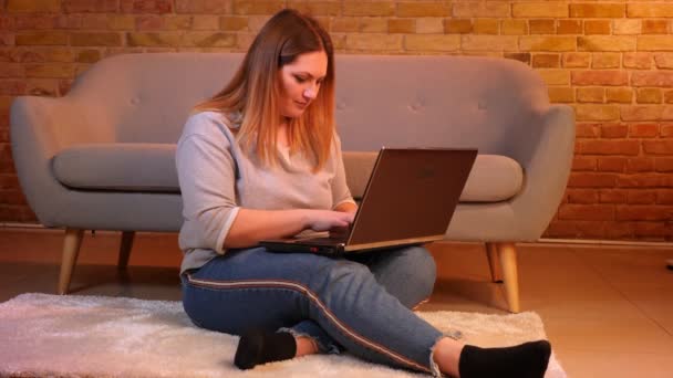 L'étudiant caucasien en surpoids est assis sur le sol en tapant attentivement sur l'ordinateur portable étant réfléchi dans une atmosphère confortable à la maison . — Video