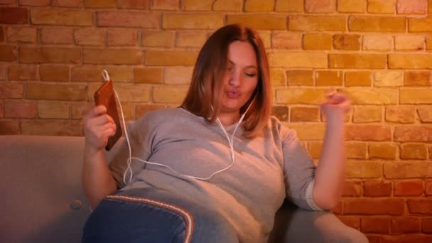 Kilolu kız öğrenci kulaklıkmüzik dinlerken ve rahat ev atmosferinde aktif dans kanepede oturur. — Stok video