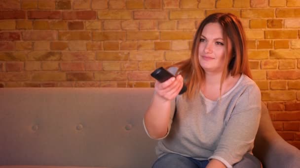 Overweight long-haired female student switches channels on TV with remote controller in cozy home atmosphere. — Stock Video