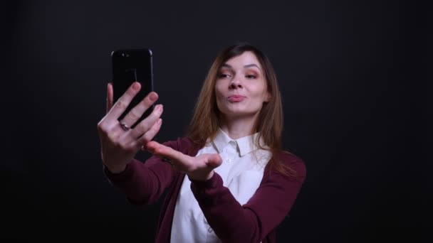 Retrato de joven mujer de negocios morena hablando en videochat en el teléfono celular alegremente sobre fondo negro . — Vídeos de Stock