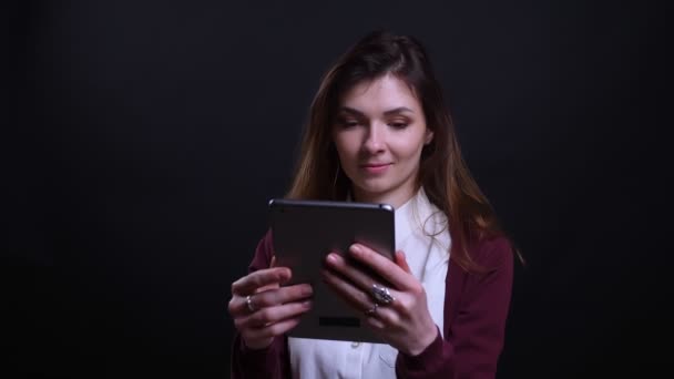 Porträt einer jungen brünetten Geschäftsfrau, die mit großem Interesse auf schwarzem Hintergrund in ein Tablet schaut.