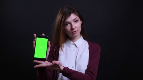 Portrait of young brunette businesswoman shows green chroma screen of phone into camera on black background. — Stock Video