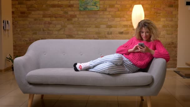 Szőke háziasszony rózsaszín pulóver pihentető kanapén szörfözés a háló segítségével okostelefon otthonos otthon hangulatát. — Stock videók