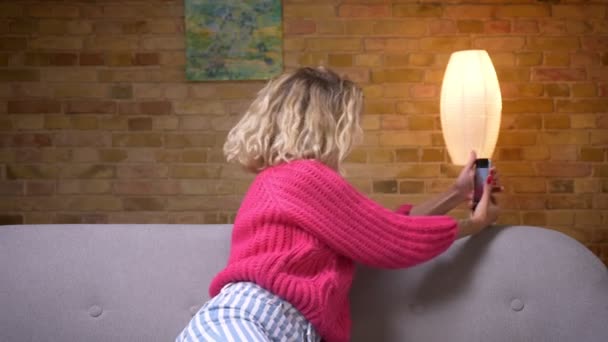 Блондинка домохозяйка в розовом свитере выбирает позицию, чтобы сделать селфи на смартфоне в уютной домашней атмосфере . — стоковое видео