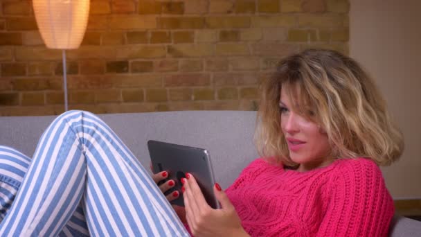 Nahaufnahme einer blonden Hausfrau im rosa Pullover, die auf dem Sofa liegt und sich im Videochat auf dem Tablet in gemütlicher häuslicher Atmosphäre unterhält. — Stockvideo
