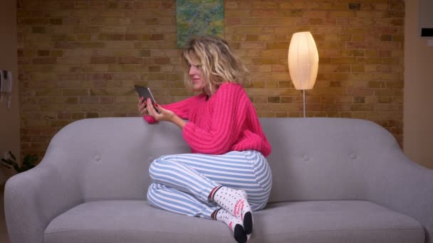 Блондинка-домохозяйка в розовом свитере сидит на диване серфинга в планшете и эмоционально реагирует в уютной домашней атмосфере . — стоковое видео