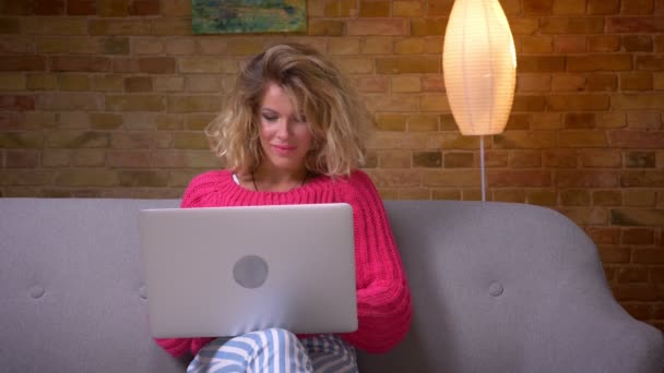 ソファの上にピンクのセーターを着た主婦は、ノートパソコンが集中して家庭の雰囲気の中で微笑んでいると注意深く働いています. — ストック動画