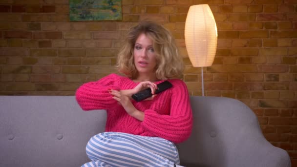 Szőke háziasszony rózsaszín pulóvert a kanapén tévénézés azt mutatja, érzelmek és ölelést magát otthonos otthon. — Stock videók