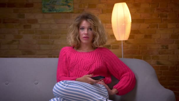 Loira dona de casa em suéter rosa no sofá assistindo TV fica aterrorizado e esconde seu rosto em casa aconchegante . — Vídeo de Stock