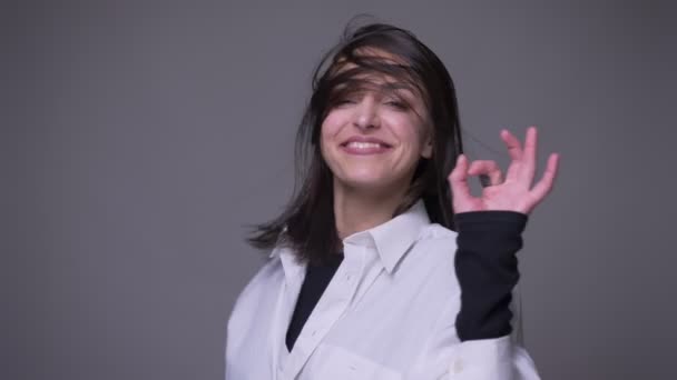 Retrato de close-up de adulto atraente mulher caucasiana girando com o cabelo tremulando e mostrando alegremente bem handsign olhando para a câmera com fundo isolado em cinza — Vídeo de Stock