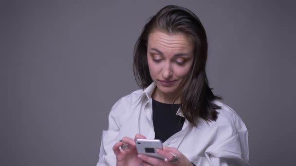 Retrato de close-up de adulto atraente caucasiano feminino mensagens de texto no telefone e sorrindo olhando para a câmera com fundo isolado em cinza — Vídeo de Stock