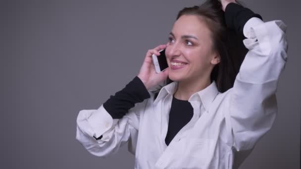 Портрет крупным планом взрослой привлекательной кавказки с телефонным звонком и улыбкой на фоне, изолированном на сером — стоковое видео