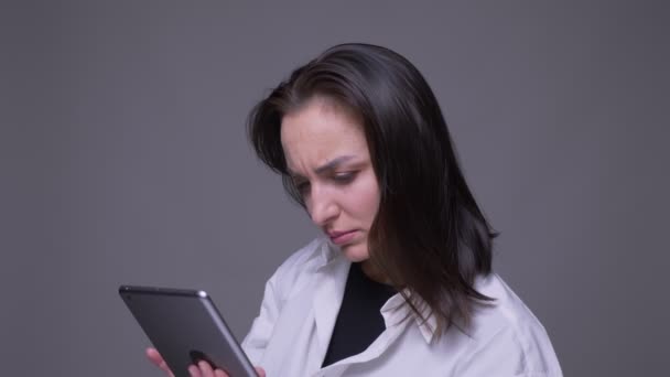 Retrato de primer plano de mensajería femenina caucásica atractiva para adultos en la tableta frente a la cámara con fondo aislado en gris — Vídeos de Stock