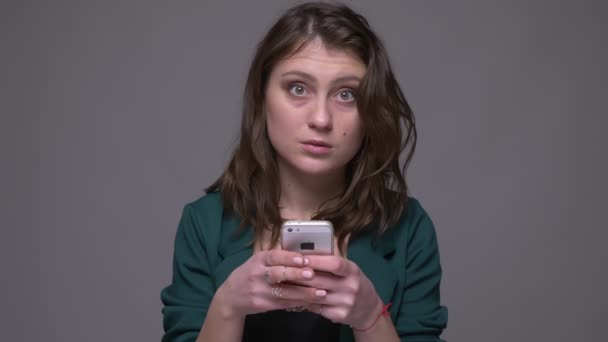 Close-up tiro de adulto atraente morena feminino usando o telefone e olhando para a câmera sorrindo com fundo isolado em cinza — Vídeo de Stock
