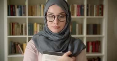 Başörtüsü ve gözlük müslüman öğrenci portresi kütüphanede elinde kitap tutan kameraya gülümsüyor.