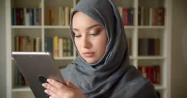 Tabletle çalışan türbanlı Müslüman öğrencinin profil portresi kütüphanede kameraya sakince bakıyor.