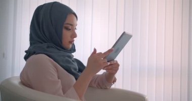Tabletle çalışan türbanlı Müslüman iş kadınının profil çekimi ofiste sakin olan kameraya dönüşüyor.