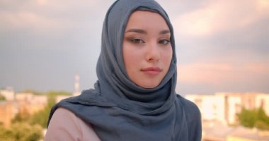 Büyük şehir manzaralı balkonda duran kameraya sakince izlerken hijab Müslüman öğrencinin yakın çekim portresi.