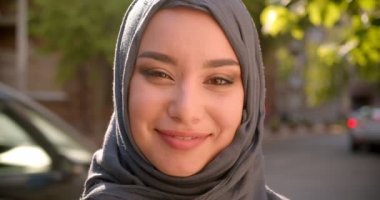 Yeşil şehir caddesinde duran kameraya neşeyle gülümseyen türbanlı Müslüman öğrenciportresi.