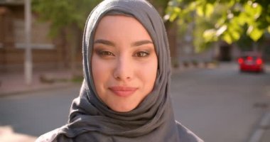 Başörtüsü müslüman öğrenci portresi gülümseyerek ve yeşil şehir caddesinde ayakta kameraya ışınlanıyor.