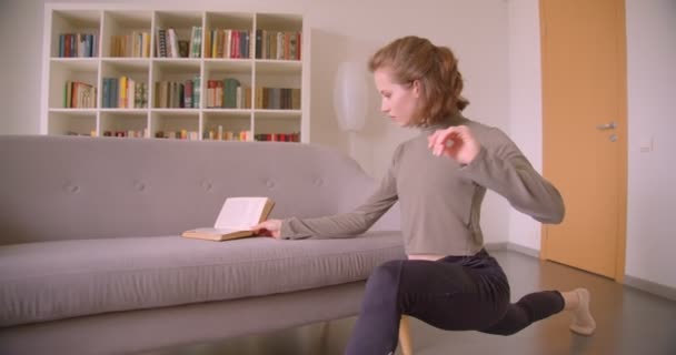 Menina caucasiana suave faz alongamento e leitura de um livro simultaneamente na sala de luz aconchegante . — Vídeo de Stock