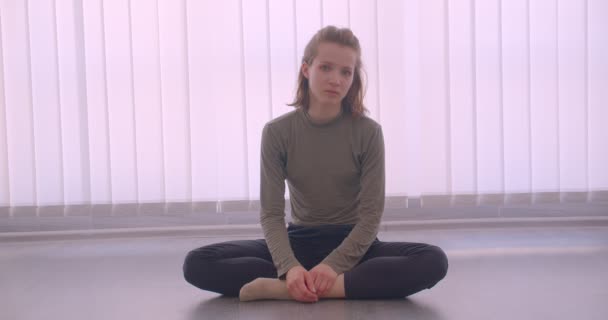 Nahaufnahme Porträt einer kaukasischen Profi-Ballerina, die im hellen Studio auf dem Boden sitzt und ruhig in die Kamera schaut. — Stockvideo