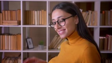 Gözlüklü esmer kadın öğretmenin kameraya mutlu bir şekilde gülümseyip kütüphanede flört eden portresi.