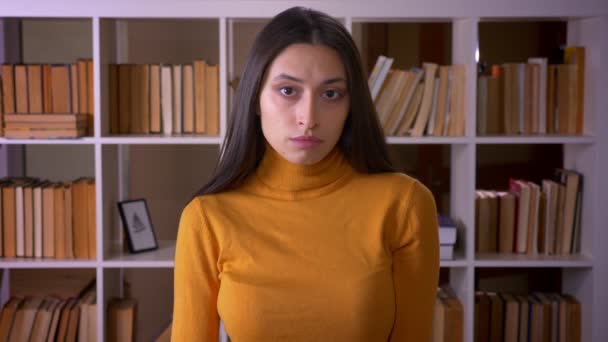 Portret van stijlvolle brunette student kijken kalm en aandachtig in de camera op de boekenkasten achtergrond. — Stockvideo