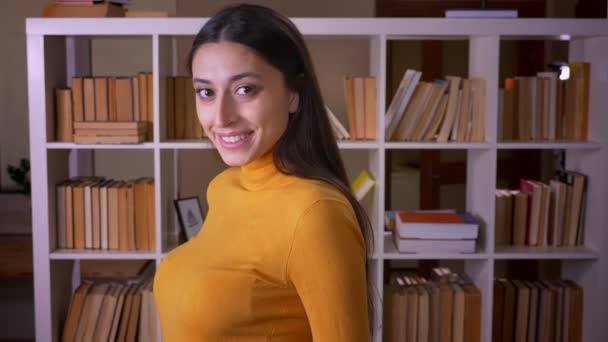 Портрет стильної брюнетки вчителька-жінка перетворюється на камеру і щасливо посміхається в неї в бібліотеці . — стокове відео