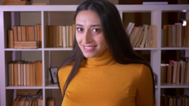 Porträtt av vacker brunett kvinnlig lärare ler blygt i kameran känner sig generad på biblioteket. — Stockvideo