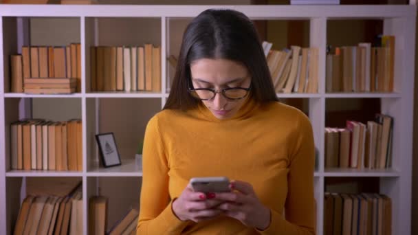 Portrait de belle enseignante brune en lunettes regardant attentivement le smartphone debout à la bibliothèque . — Video