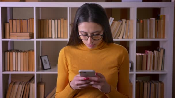 Портрет красивой брюнетки учительницы в очках, смотрящей в смартфон, внимательно стоящей в библиотеке . — стоковое видео